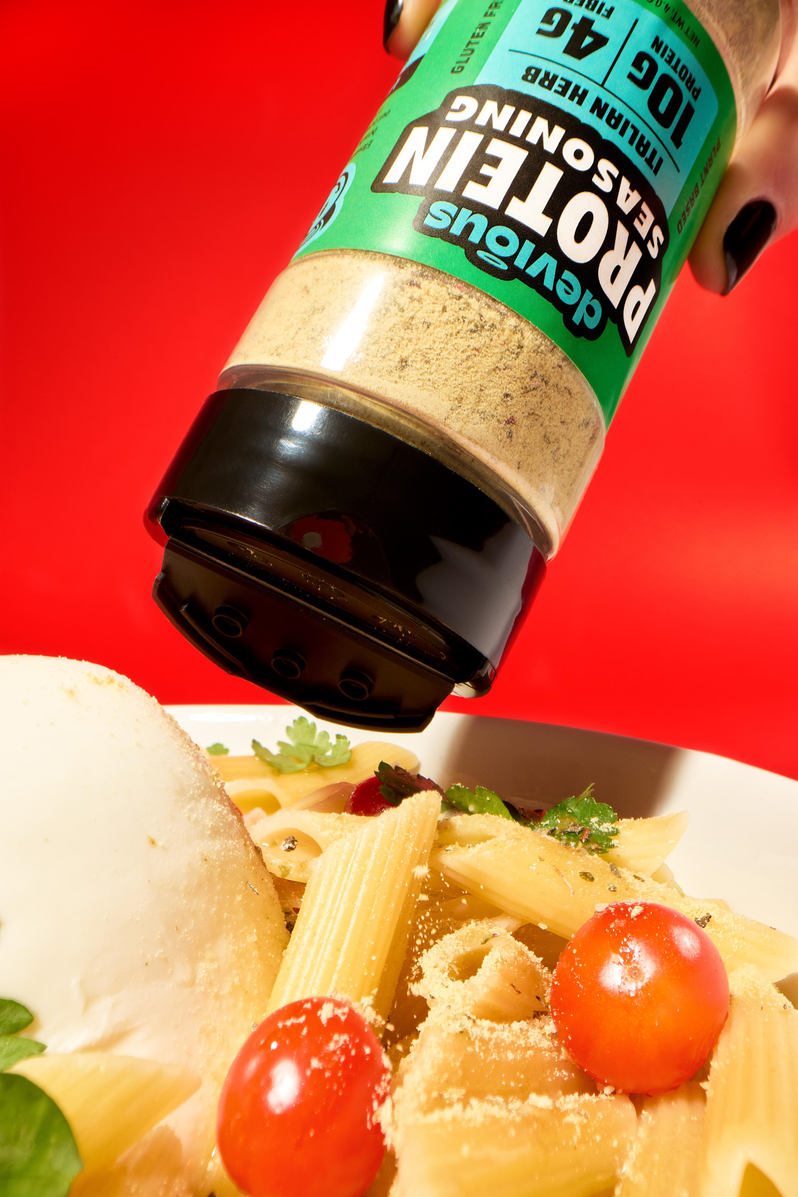 Pasta dish with protein seasoning being sprinkled on it against a red background