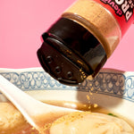 Bottle of protein seasoning being sprinkled into a bowl of soup with dumplings on a pink background
