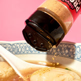 Bottle of protein seasoning being sprinkled into a bowl of soup with dumplings on a pink background