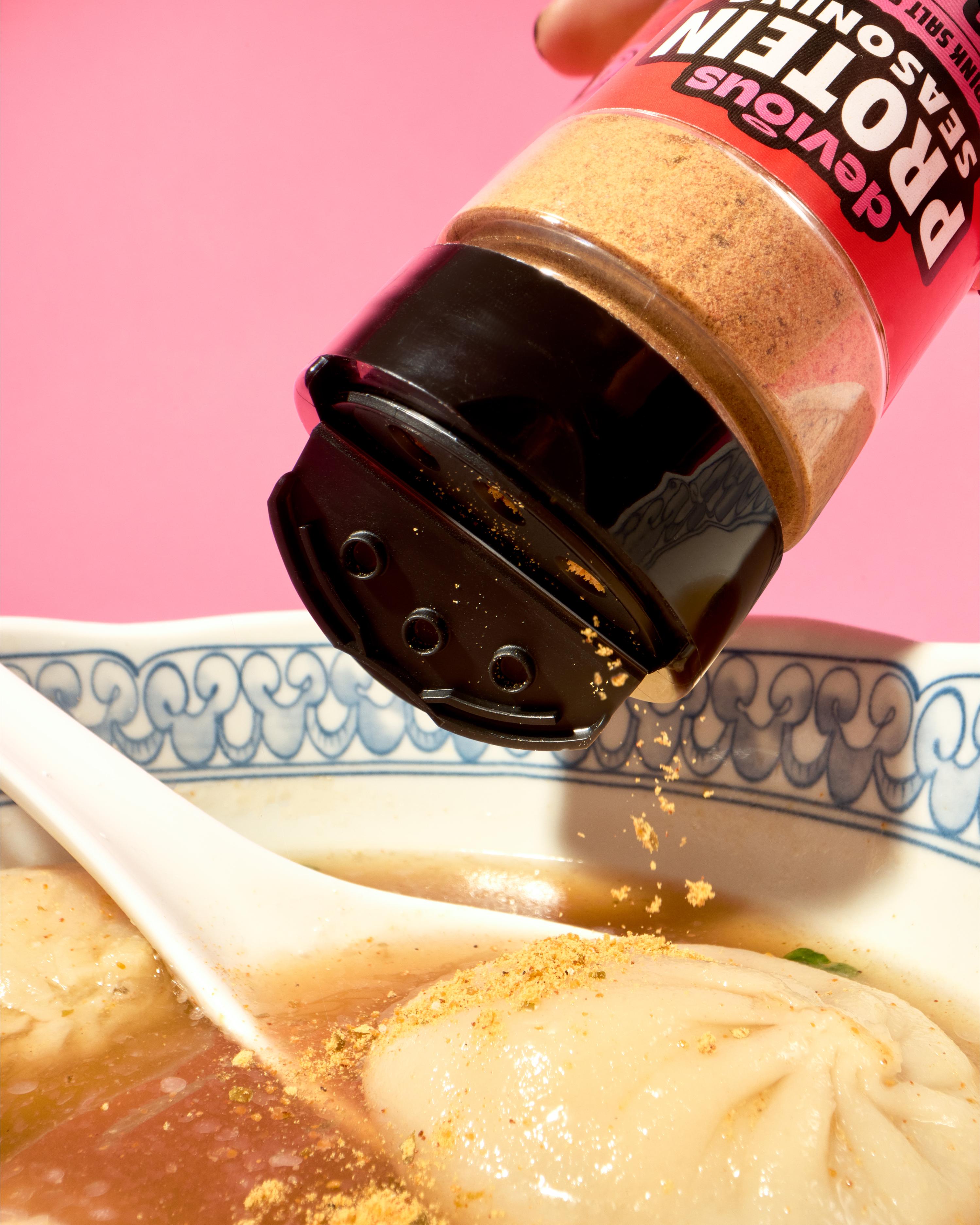 Bottle of protein seasoning being sprinkled into a bowl of soup with dumplings on a pink background