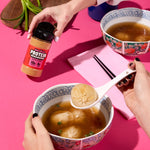 Person holding a bowl of soup with dumplings, using a spoon, on a pink background.
