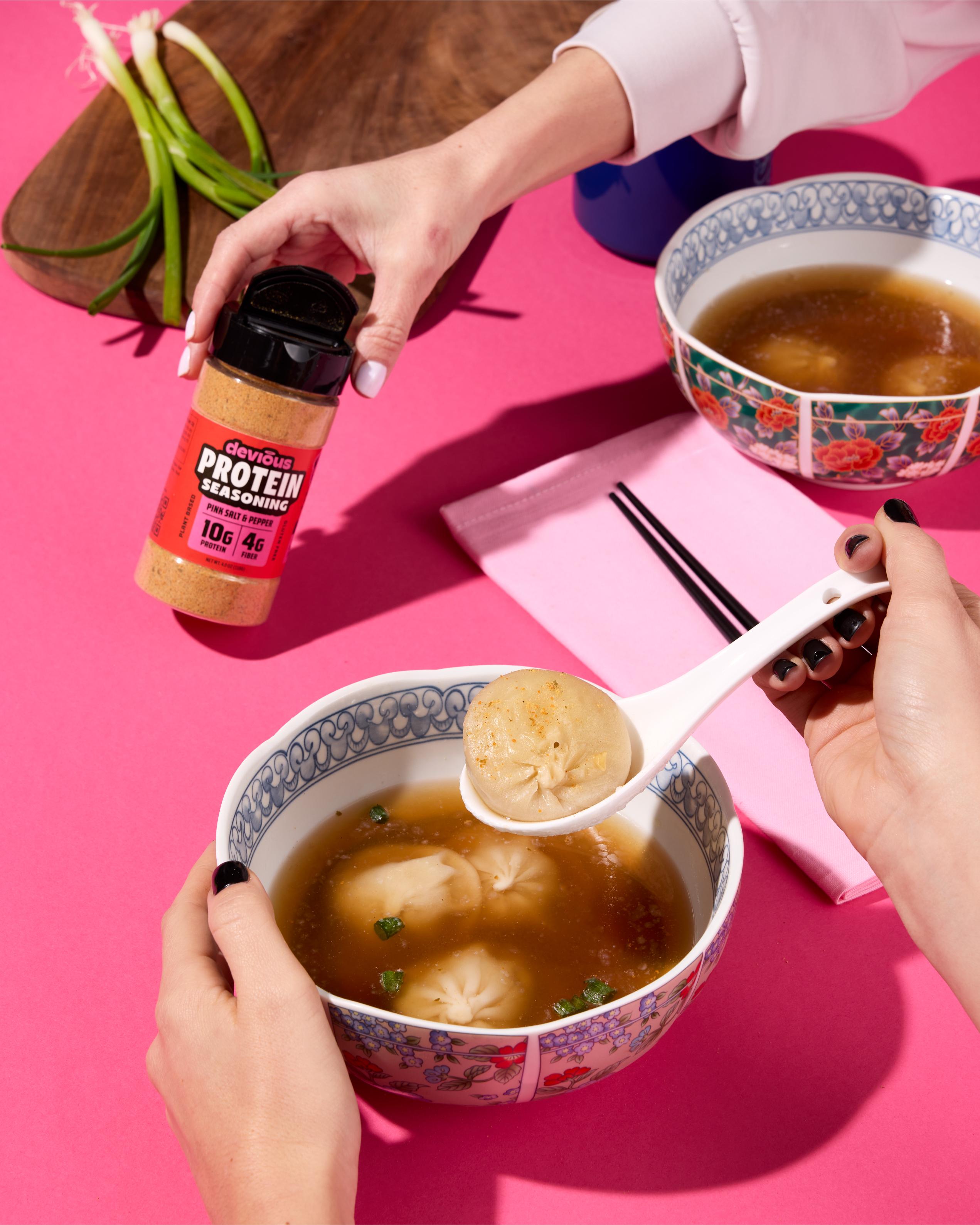 Person holding a bowl of soup with dumplings, using a spoon, on a pink background.